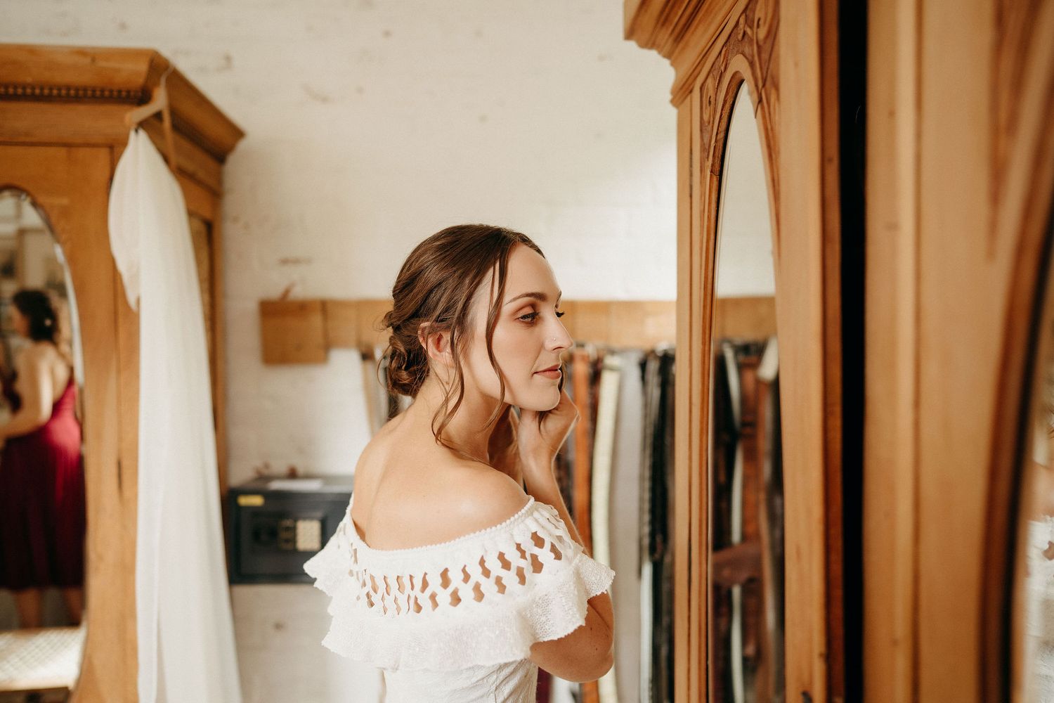 A romantic off-shoulder lace dress is reflected in an ornate wooden mirror frame in a warm-lit dressing room.