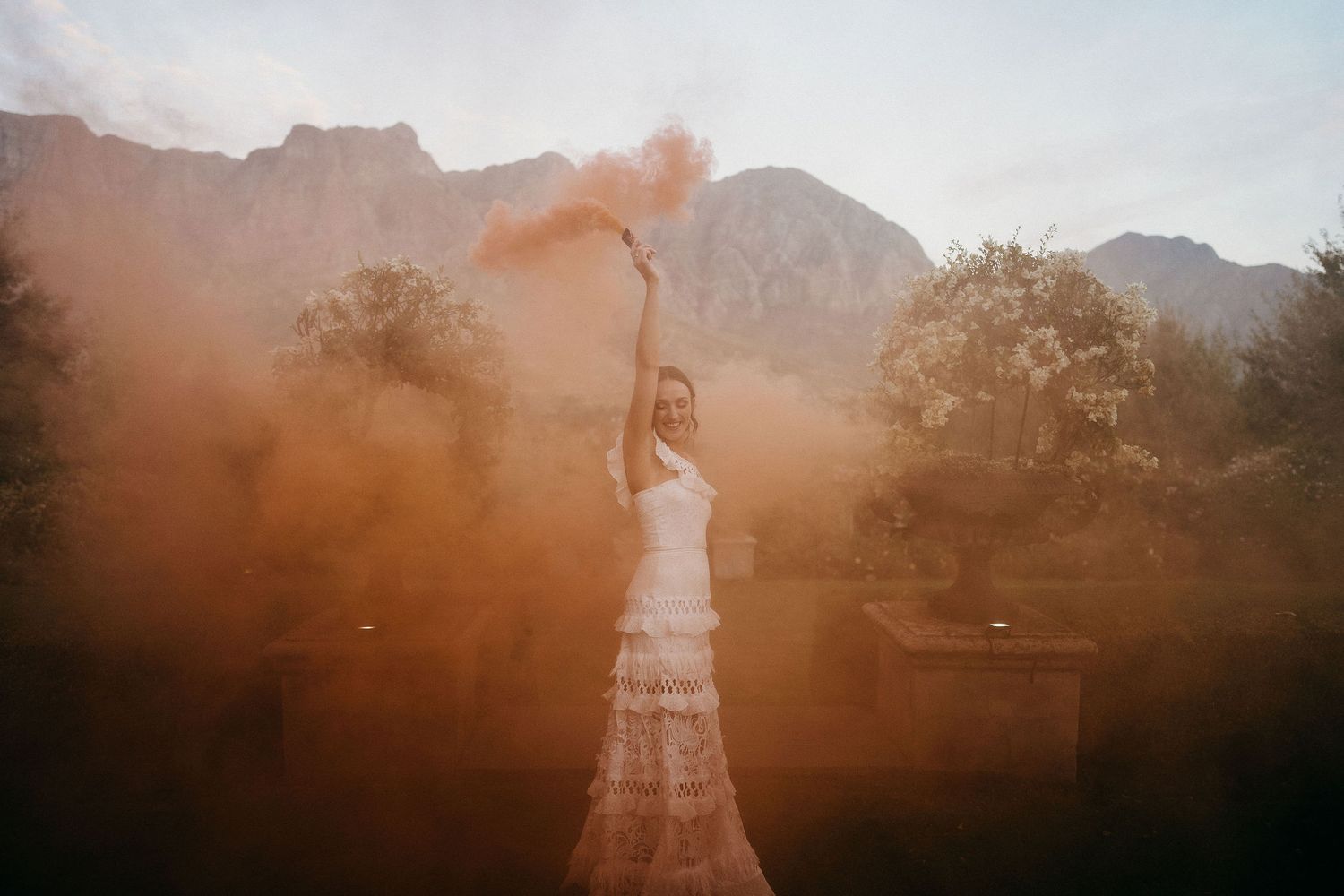 A person in a lace dress holds a smoke flare creating an ethereal orange glow against a mountain backdrop at sunset.
