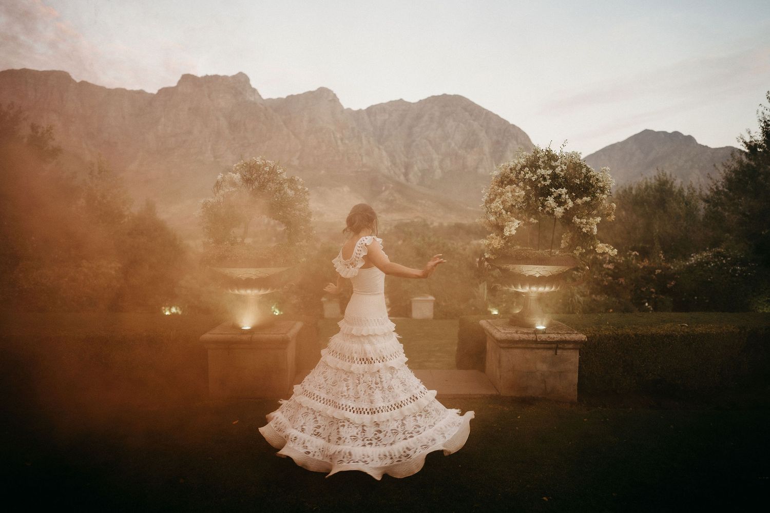 A figure in a white wedding dress twirls at sunset with dramatic mountain peaks in the background.