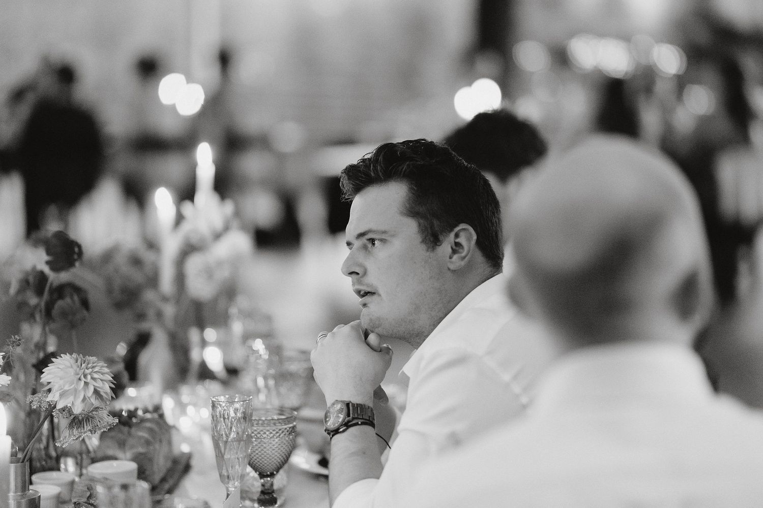 A contemplative guest in a white shirt sits at an elegant dinner table with candles and flowers in black and white.