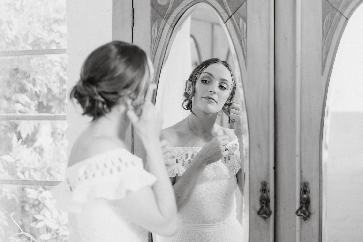 A bride in a white lace gown applies her jewelry while looking in an elegant vintage mirror.