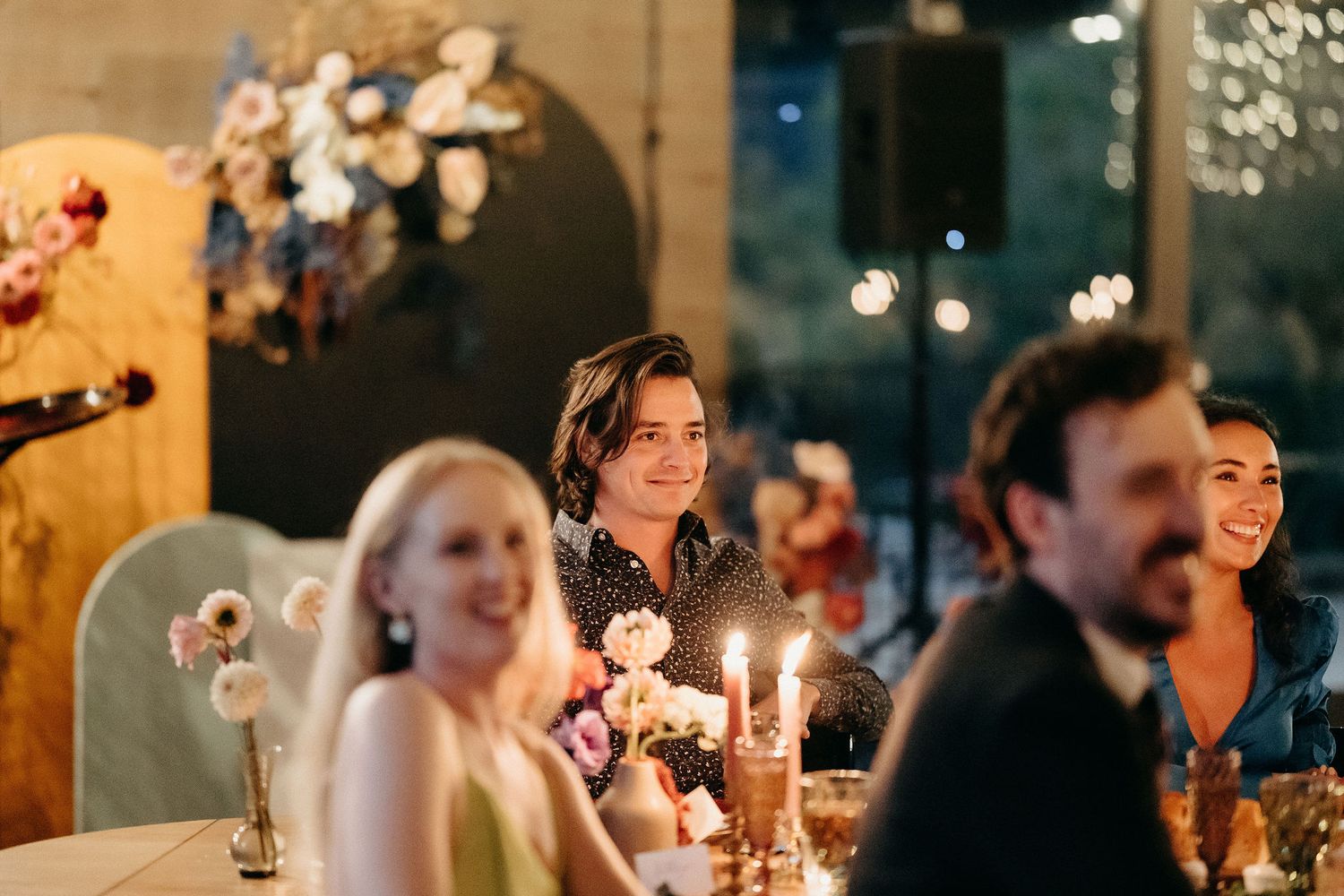 Guests enjoy an elegant candlelit dinner party with glowing lights and decorative floral arrangements in the background.