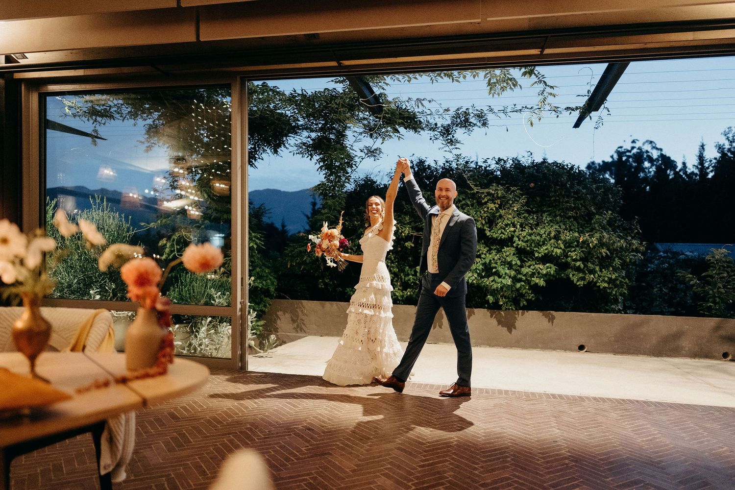 A couple dances together on a covered patio with a scenic mountain view at sunset during their wedding celebration.
