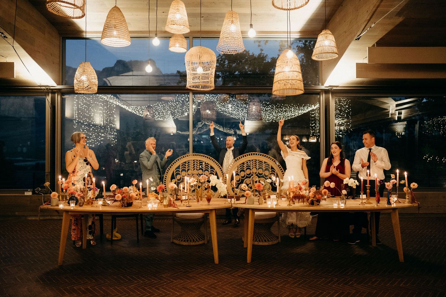 Evening wedding reception dinner under woven pendant lights with guests seated at a long rustic wooden table overlooking water.