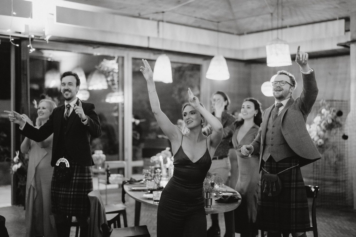 A black and white photograph showing people with raised arms dancing and celebrating in what appears to be a bar or social venue.