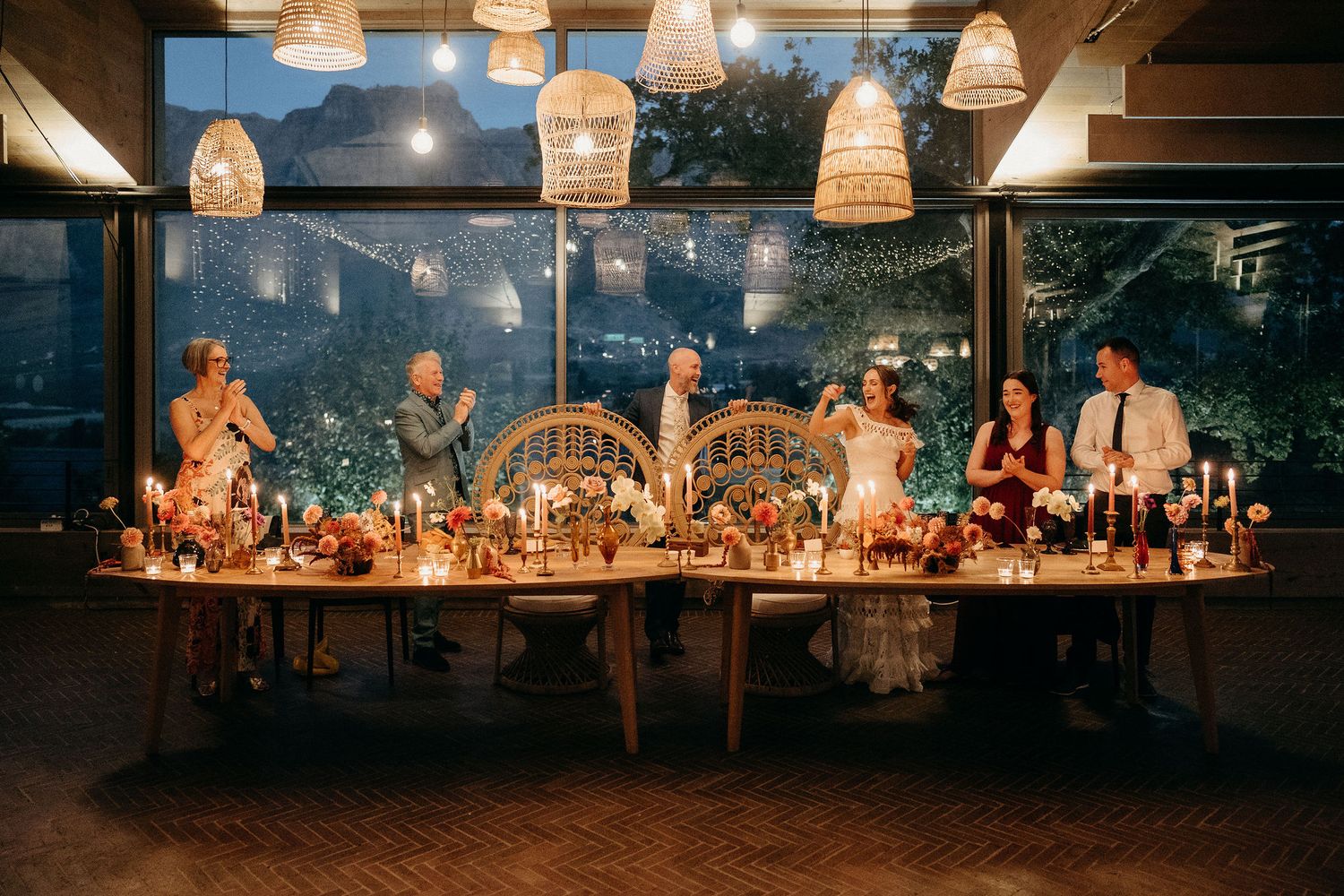 Wedding party seated at an elegantly decorated dining table under hanging wicker lamps at dusk.