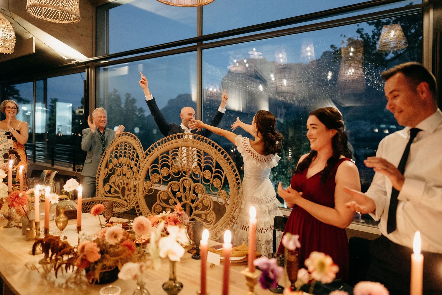 Wedding reception guests gathered at an elegantly decorated candlelit dinner table with large windows overlooking a night view.