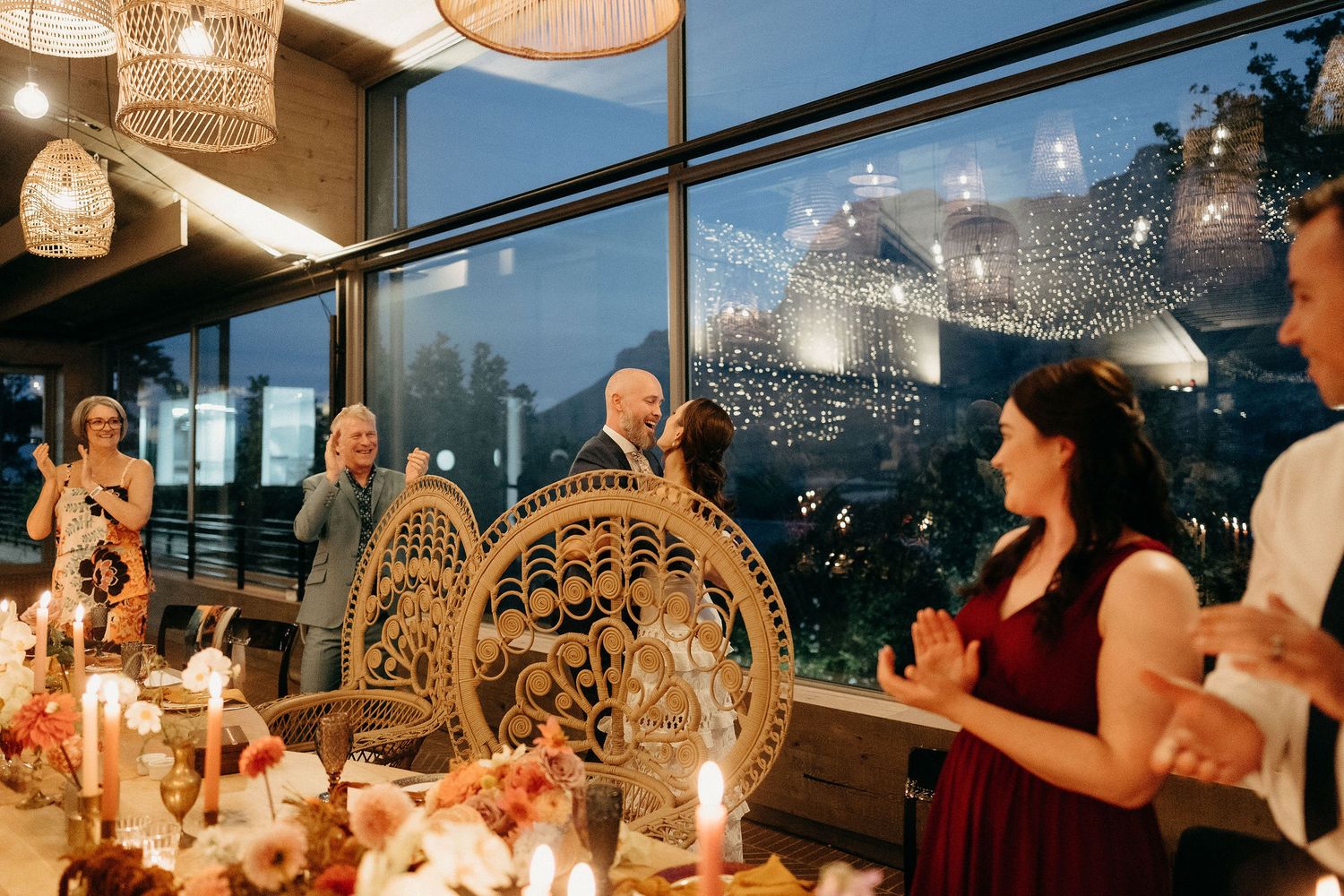 Elegant reception dinner featuring peacock chairs and twinkling lights by large windows at dusk.
