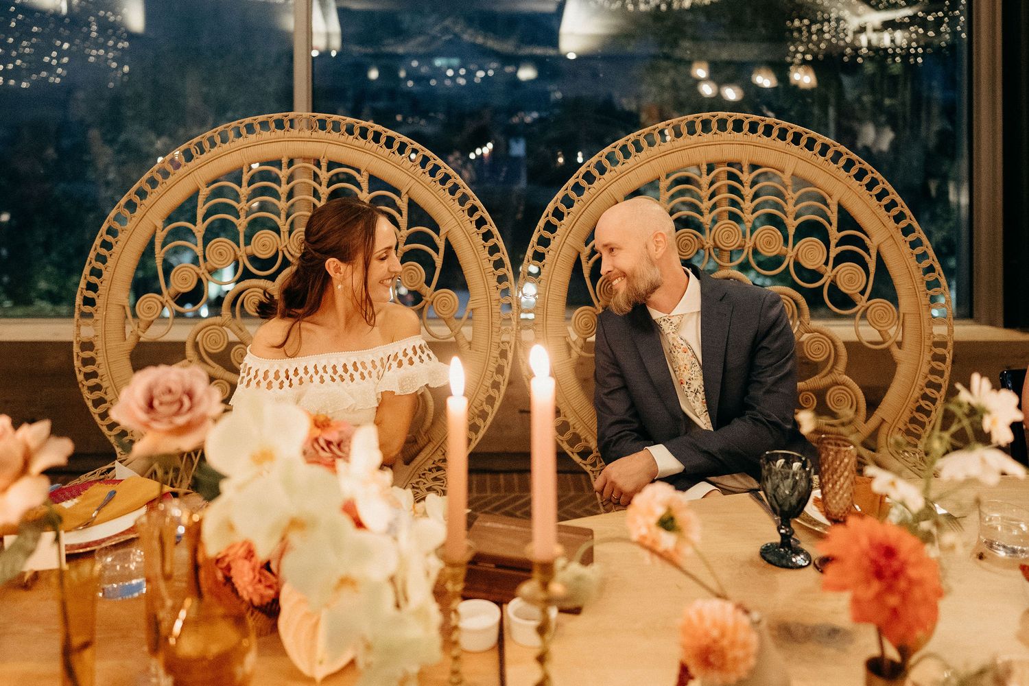 A wedding reception table setting with ornate peacock chairs, floral centerpieces and candles lit in a romantic evening setting.