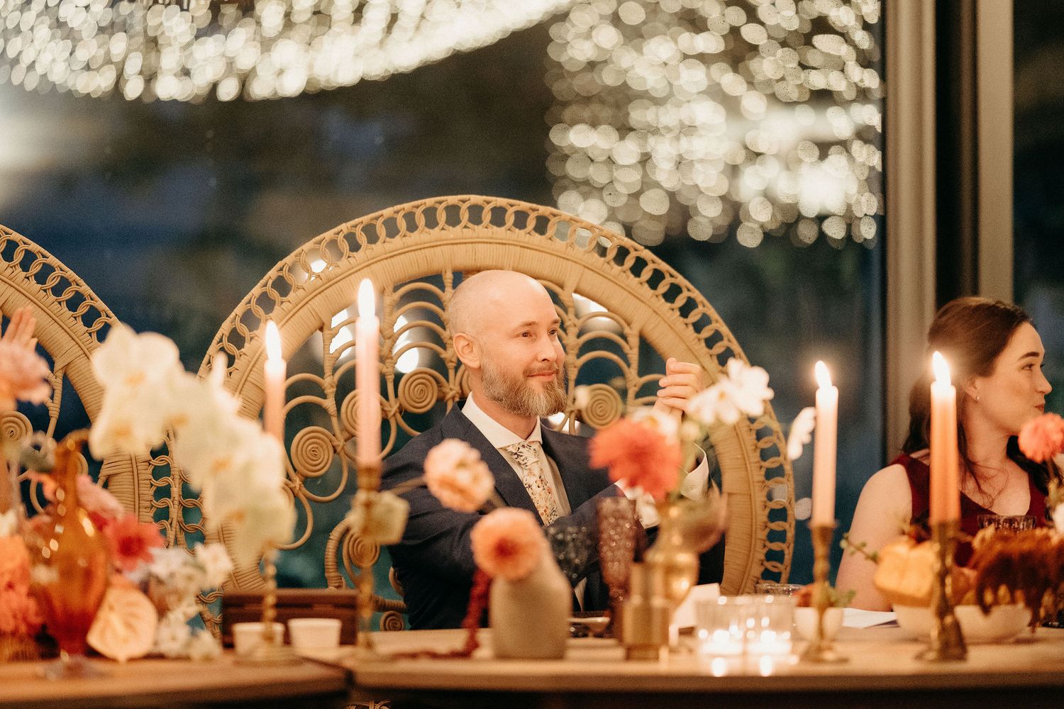 Wedding guests laugh and smile at an elegant candlelit reception dinner table with decorative chairs and warm lighting.