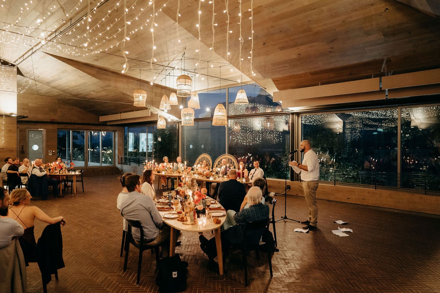 Wedding reception venue with string lights and wooden beams featuring round tables set for dinner in warm lighting.