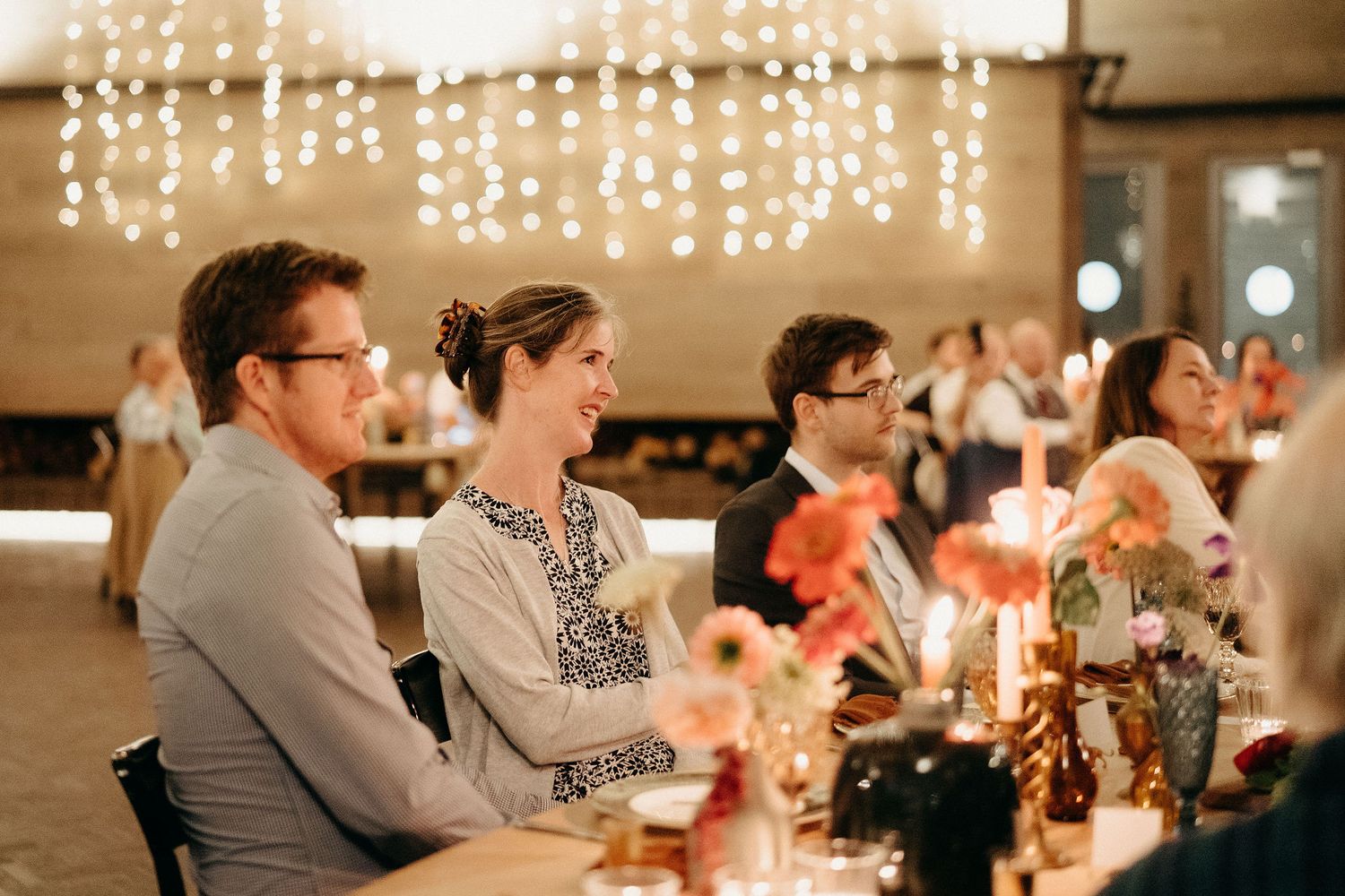 Wedding reception guests sit at a long table decorated with candles and floral arrangements under string lights.