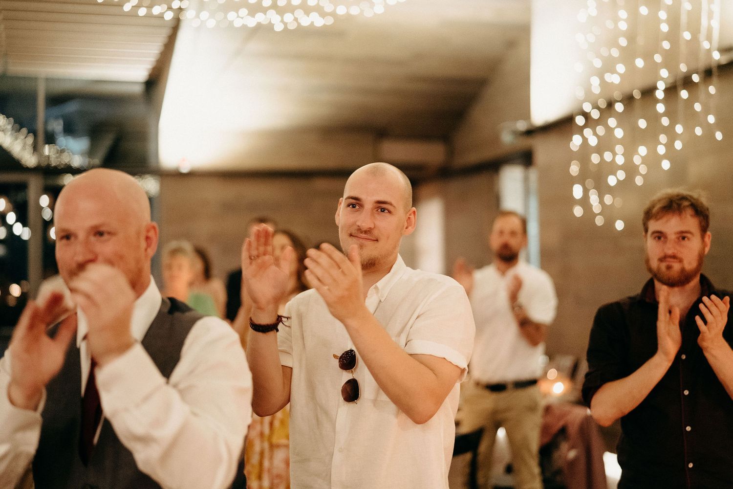 People clapping and cheering at an indoor celebration event with string lights illuminating the warm atmosphere.
