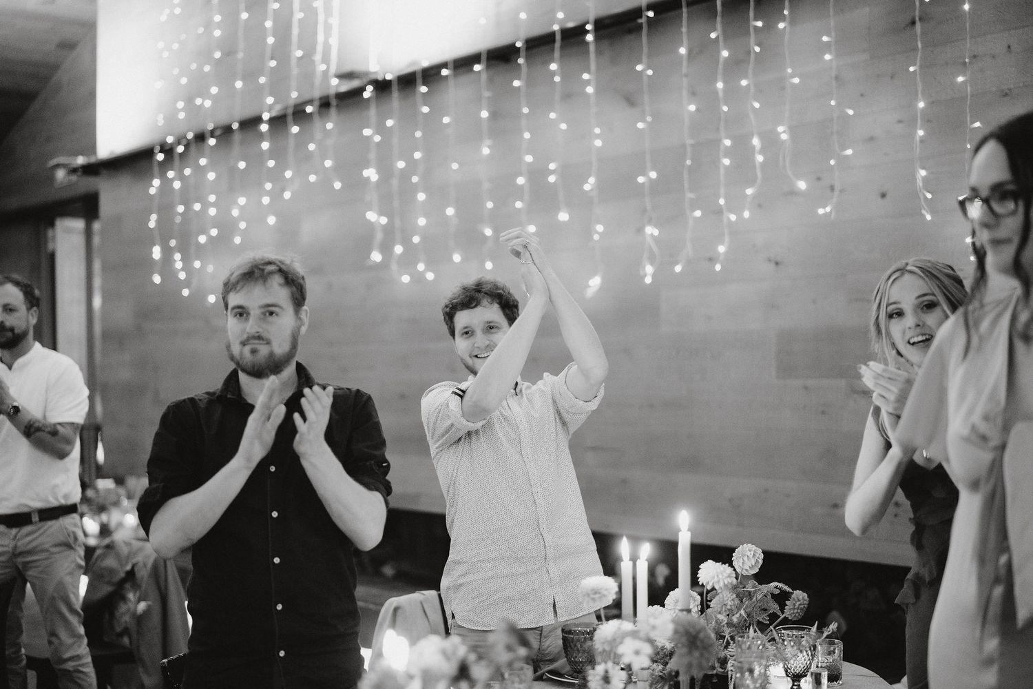 A black and white wedding reception photo with falling string lights and celebratory gathering at tables.