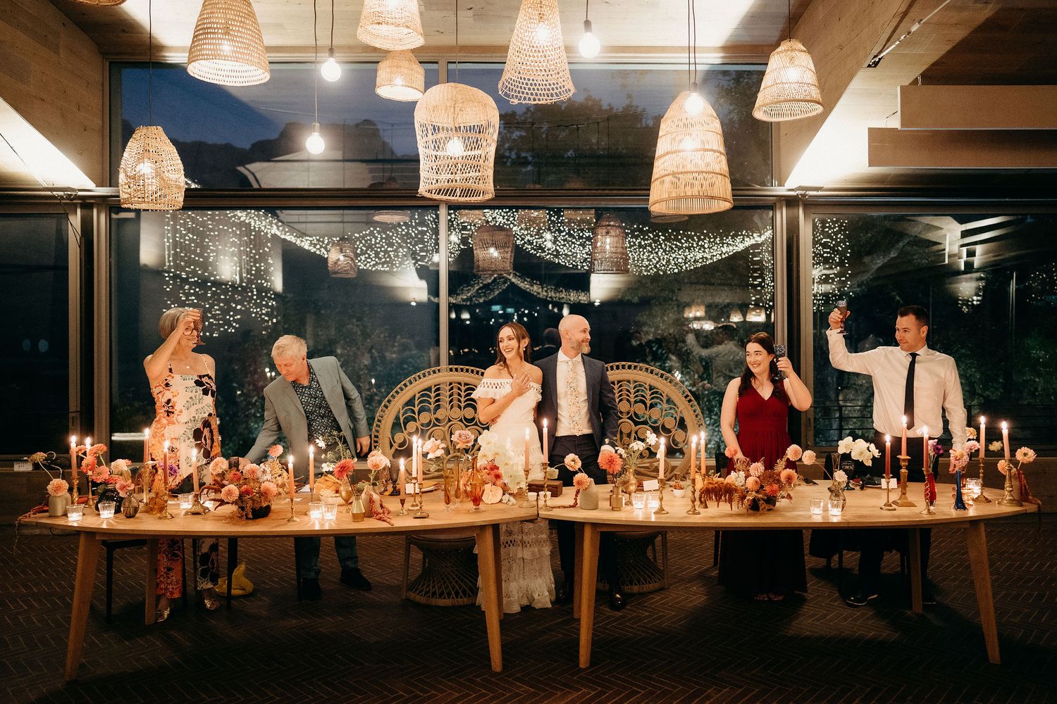 A candlelit dinner party table with guests seated under woven pendant lights and blue evening sky outside.