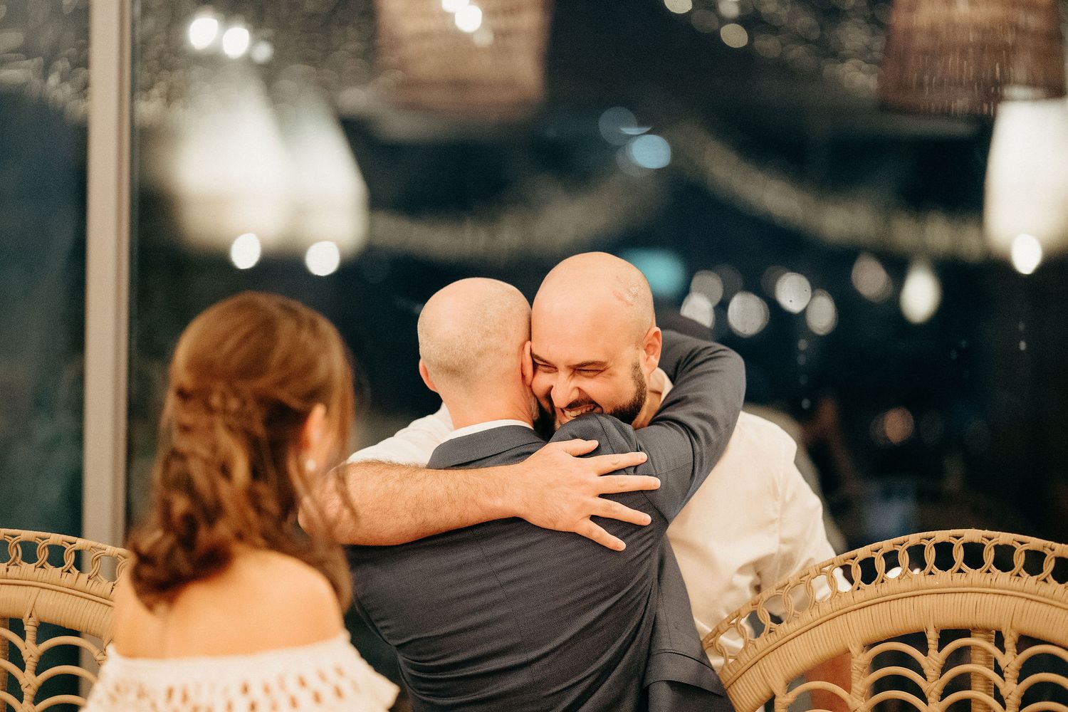 Two people embrace in a heartfelt hug during an evening celebration with bokeh lights and decorative details in the background.