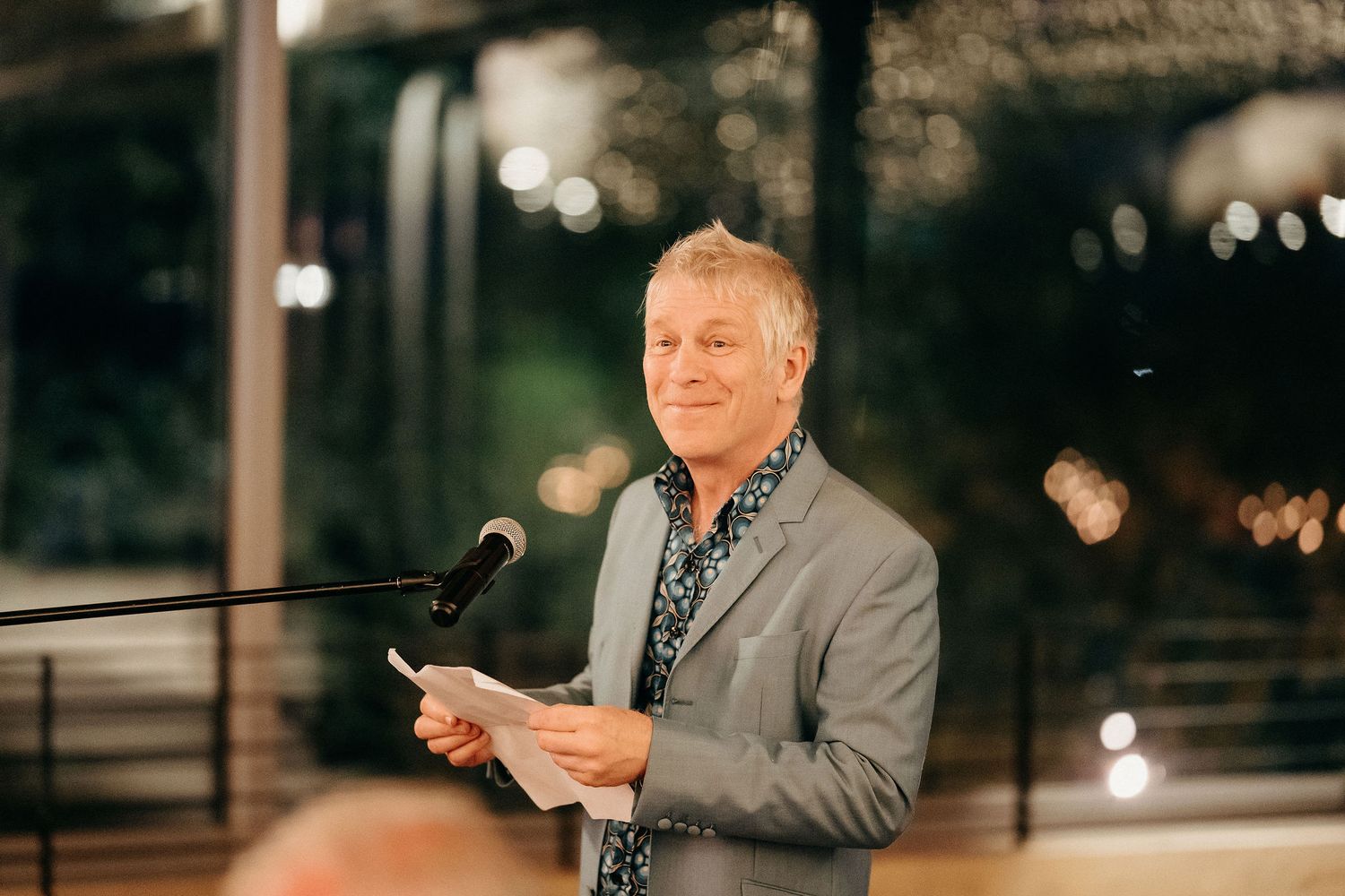 A speaker in a gray suit jacket reads from papers at a podium during an evening event.