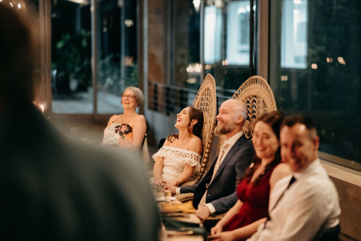 Wedding guests laugh while listening to speeches during an indoor evening reception dinner.