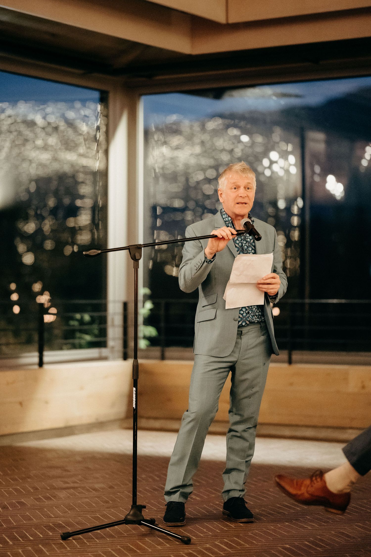 A person in a gray suit stands at a microphone reading from papers during an evening event with bokeh lights in background.