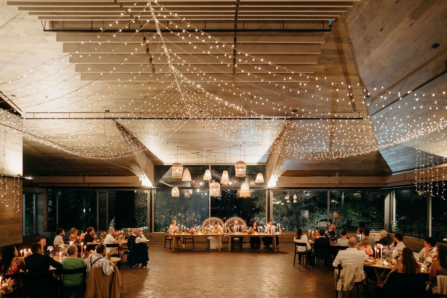 Reception hall decorated with string lights and pendant lamps hosting indoor wedding celebration with guests at dinner tables.