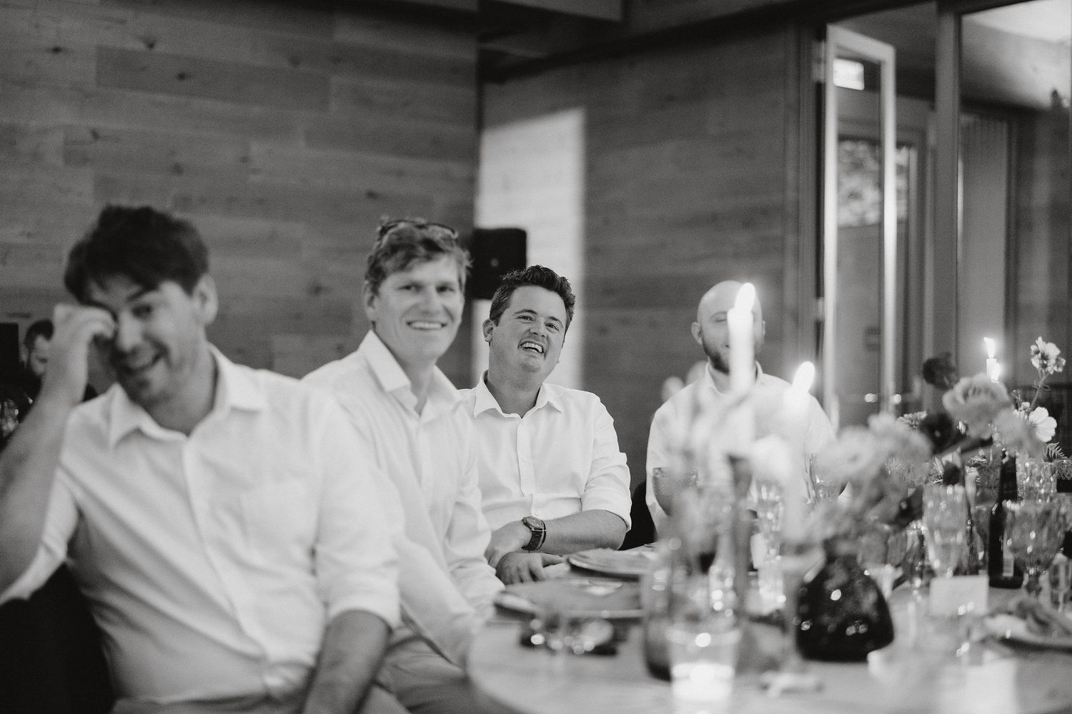 Black and white photo of guests laughing together at a dinner table with flowers and place settings.