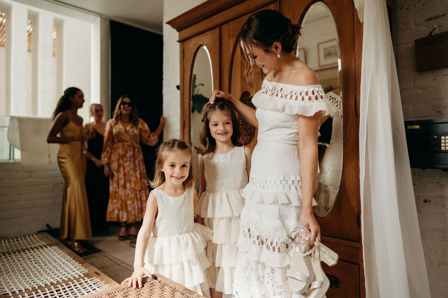 People wearing elegant white lacy dresses stand in a room with rustic furnishings during a special occasion.