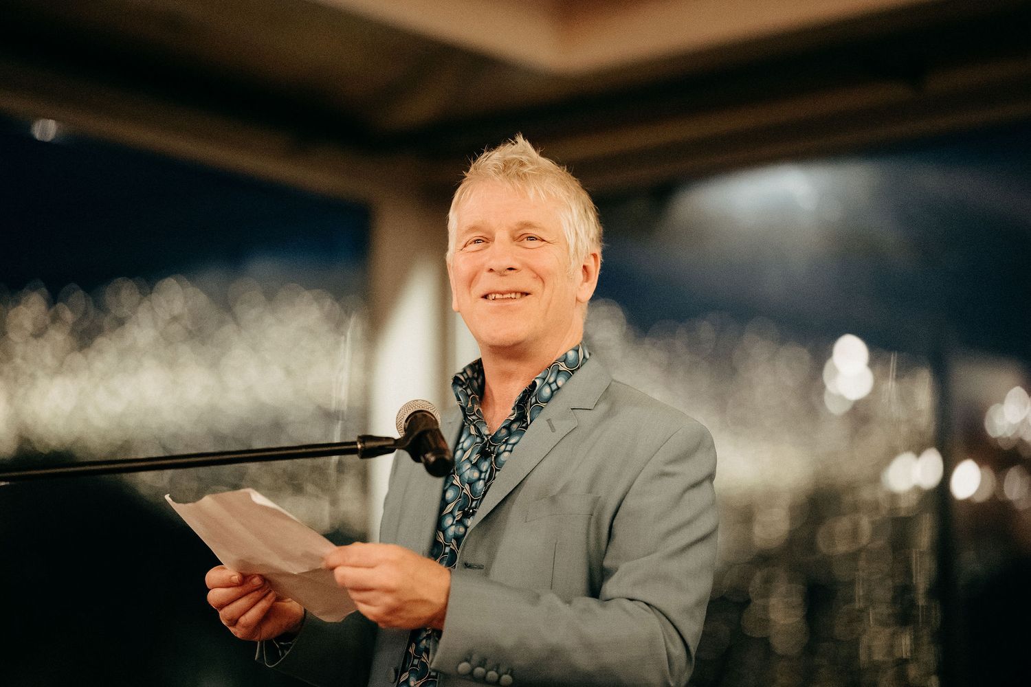 A speaker stands at a podium with a tablet device in hand, smiling under warm lighting during a presentation.