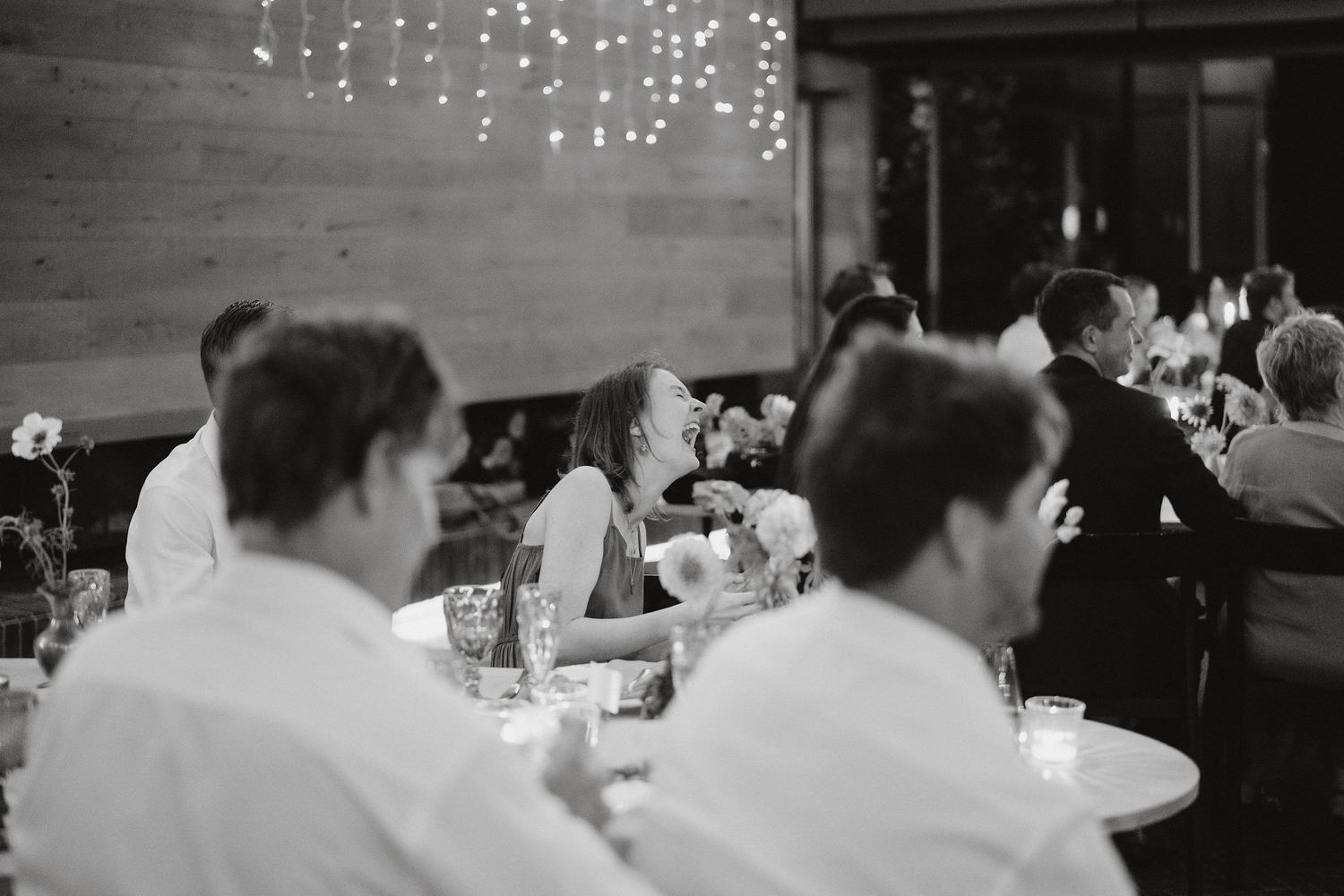 A black and white photograph showing people dining at tables in a restaurant with string lights on the wall.