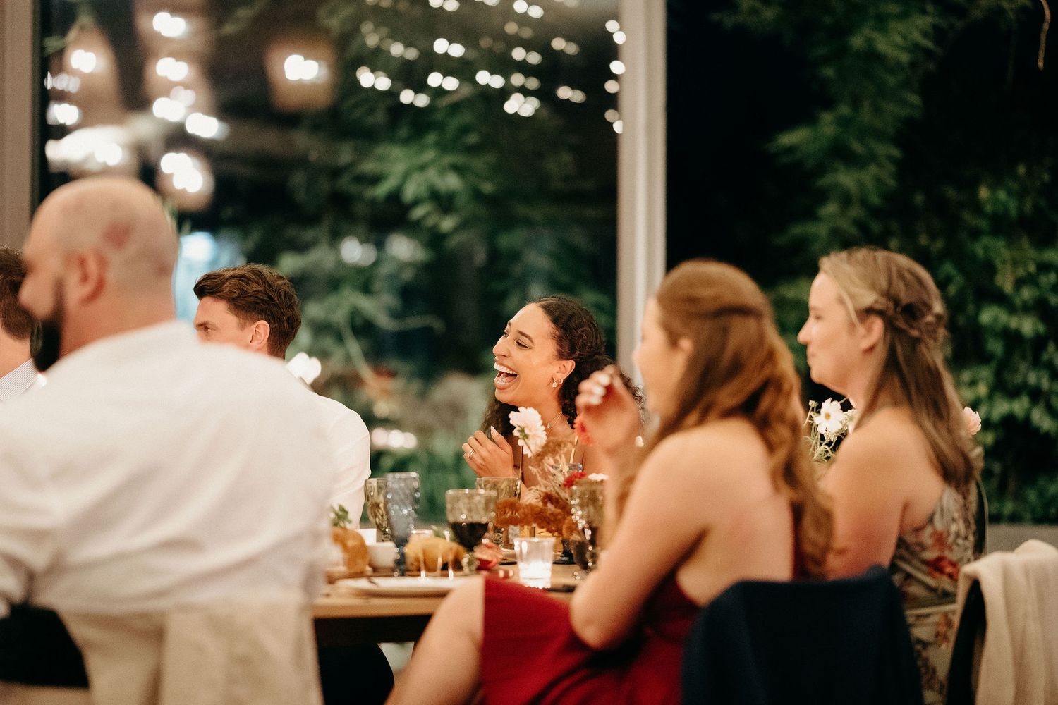 Guests enjoy an evening celebration at a reception with warm lighting and laughter around dinner tables.