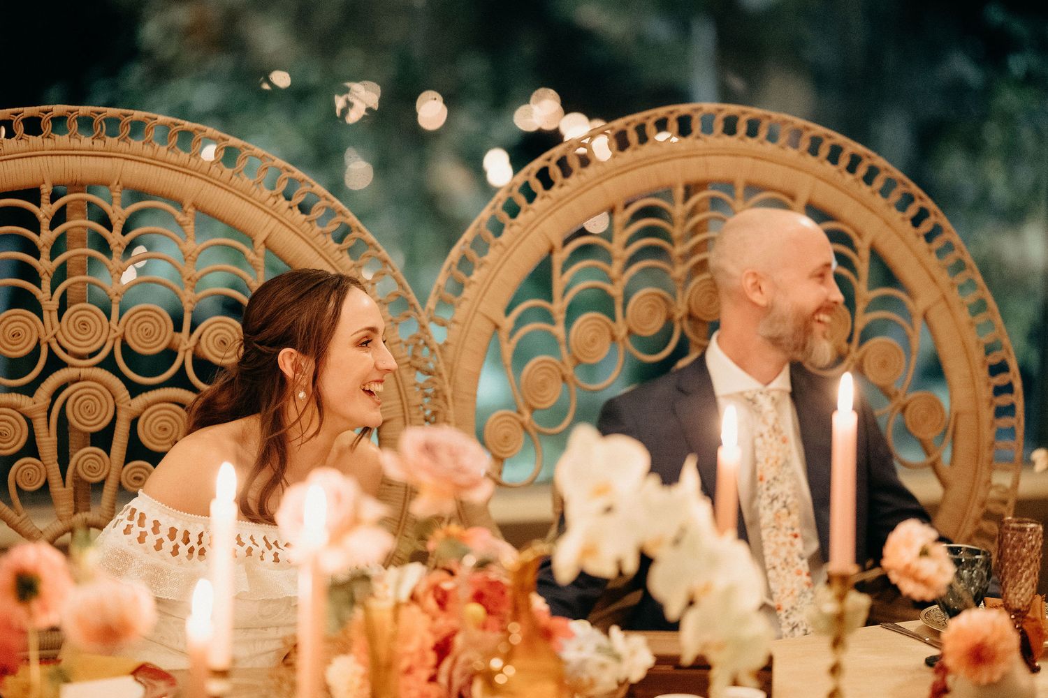 A couple sits at an elegant wedding reception table decorated with orchids and ornate peacock chairs.