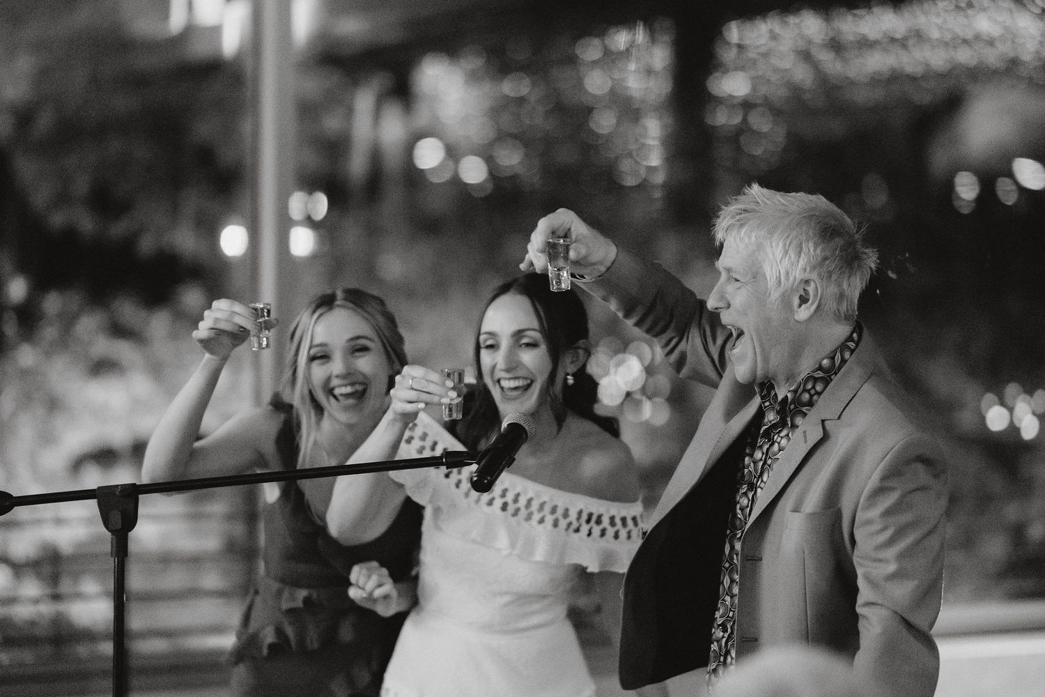 A candid black and white photo of joyful wedding party celebration with raised arms and laughter at an outdoor reception.