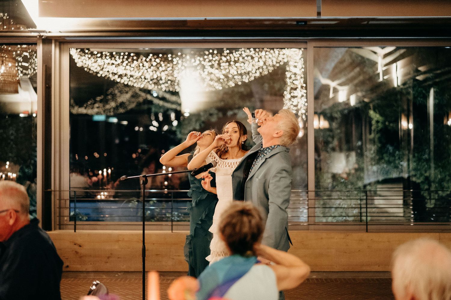 A couple dances intimately beneath twinkling lights at an indoor venue while guests look on from their seats.