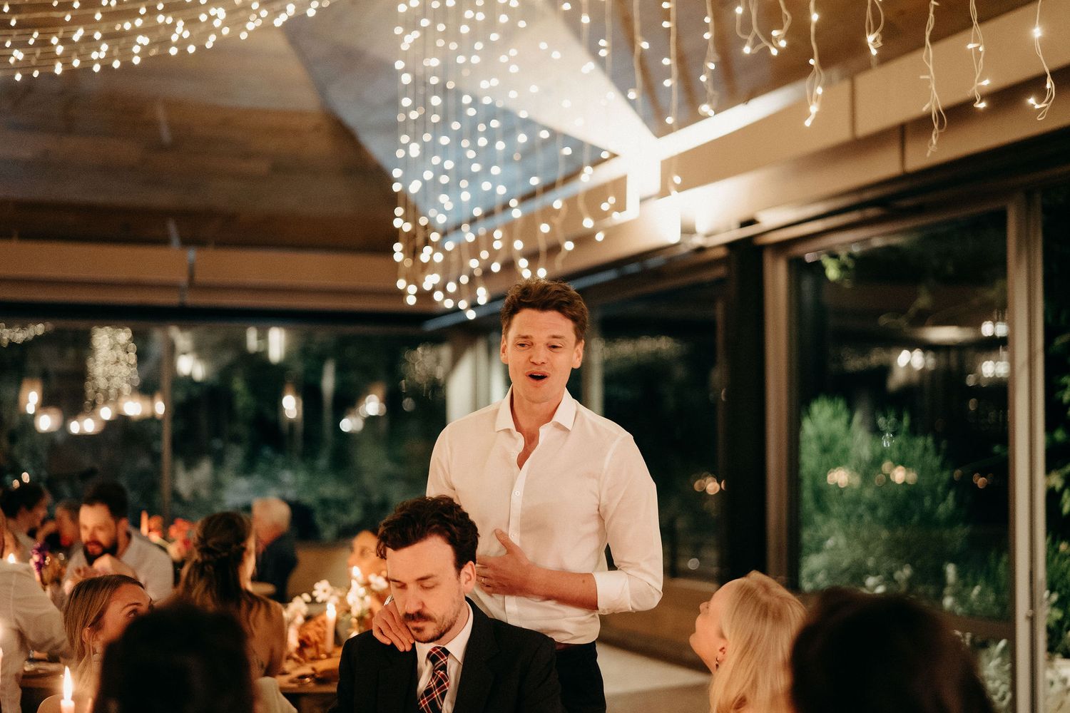 People enjoying a festive dinner gathering under sparkling string lights at an elegant indoor-outdoor venue.