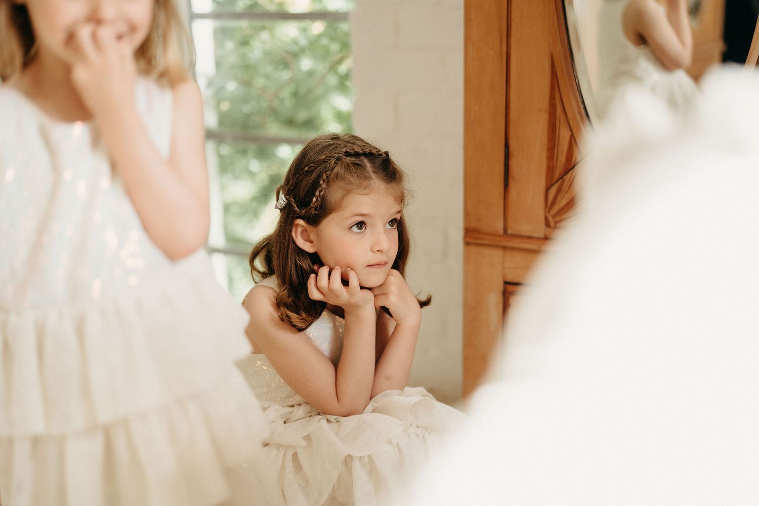 A flower girl in a white dress rests her chin on her hands with a dreamy expression.
