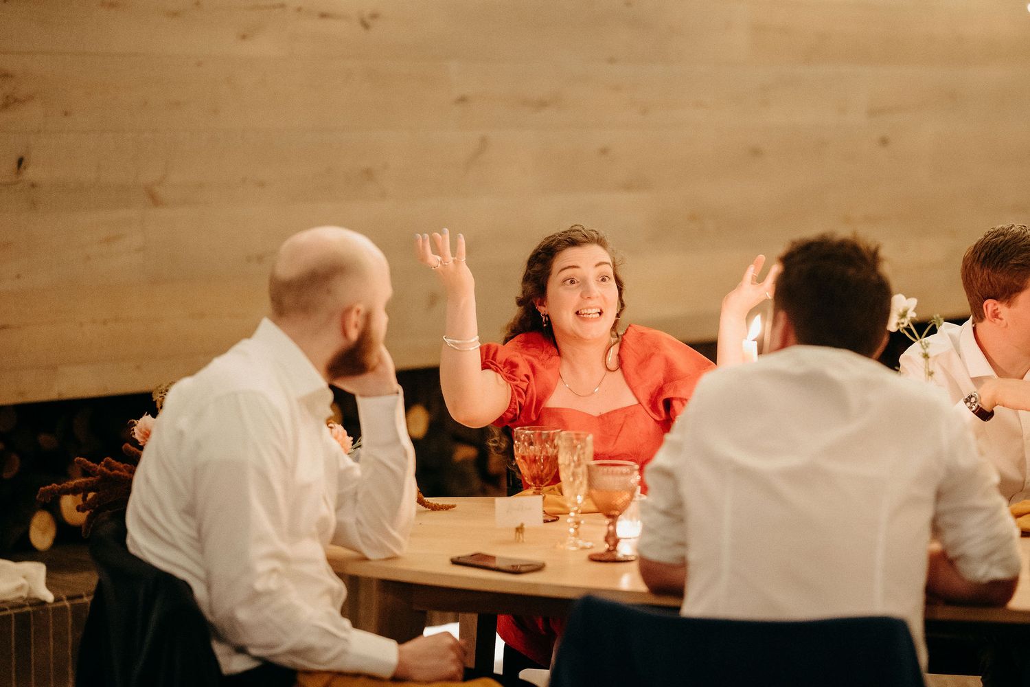 Group of friends enjoying animated conversation over drinks at a restaurant table with wooden wall backdrop.