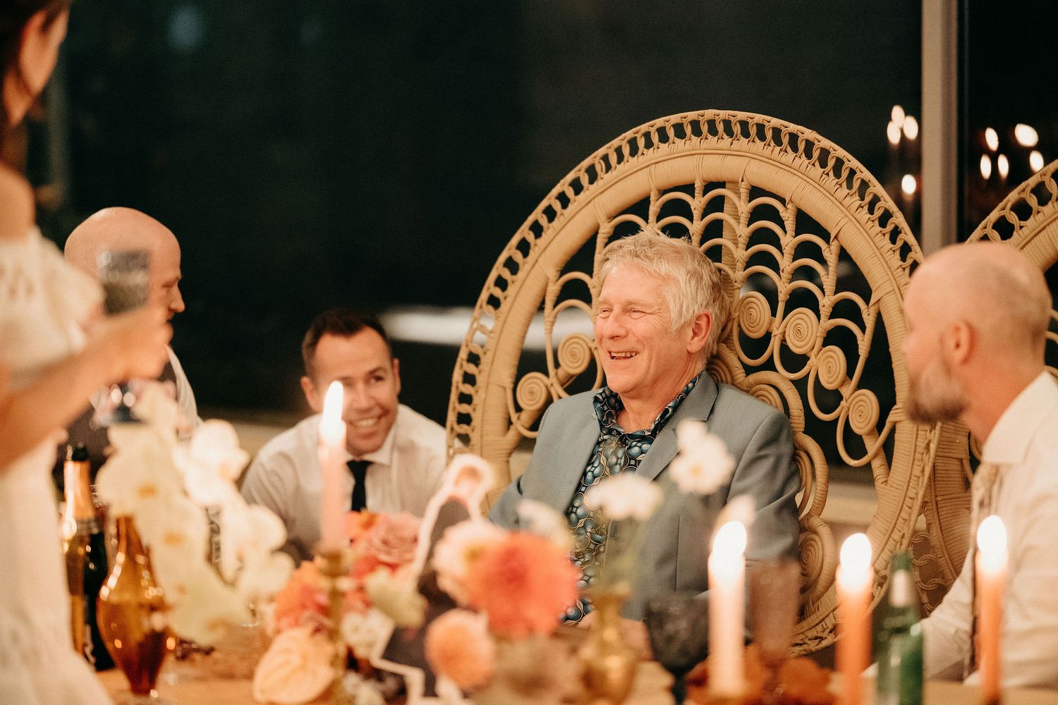 People enjoying conversation and laughter at an elegant dinner table decorated with flowers and a woven rattan chair.