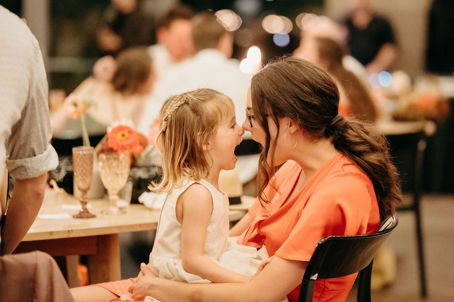 Two people share a tender moment at a dinner table with bokeh lighting and table settings in the background.