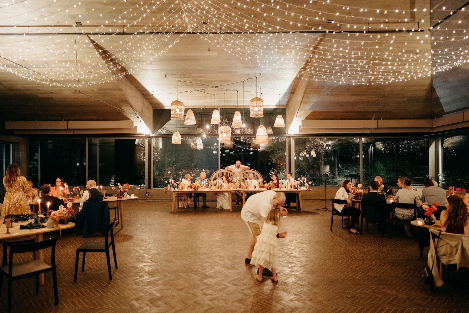 Couple dances under twinkling string lights at a rustic indoor reception venue with guests seated at surrounding tables.