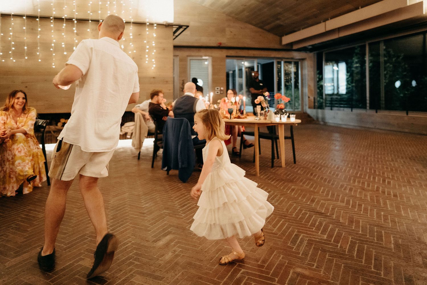 People dancing at a wedding reception in an indoor venue with string lights and herringbone wood flooring.
