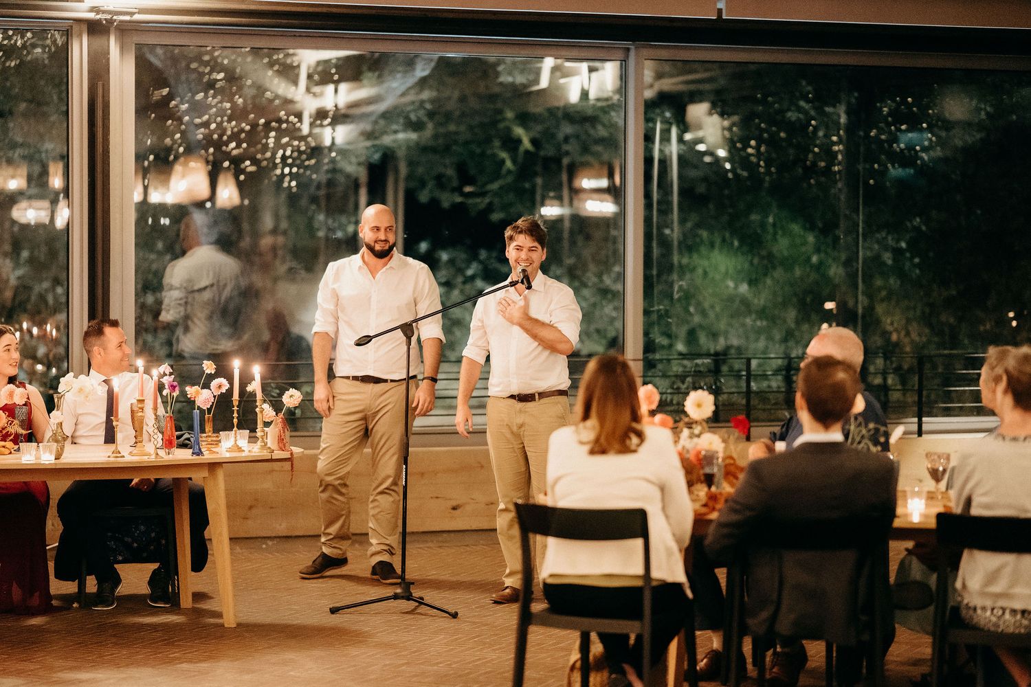 During a wedding reception, speakers share words while guests listen in a modern glass-enclosed venue at sunset.