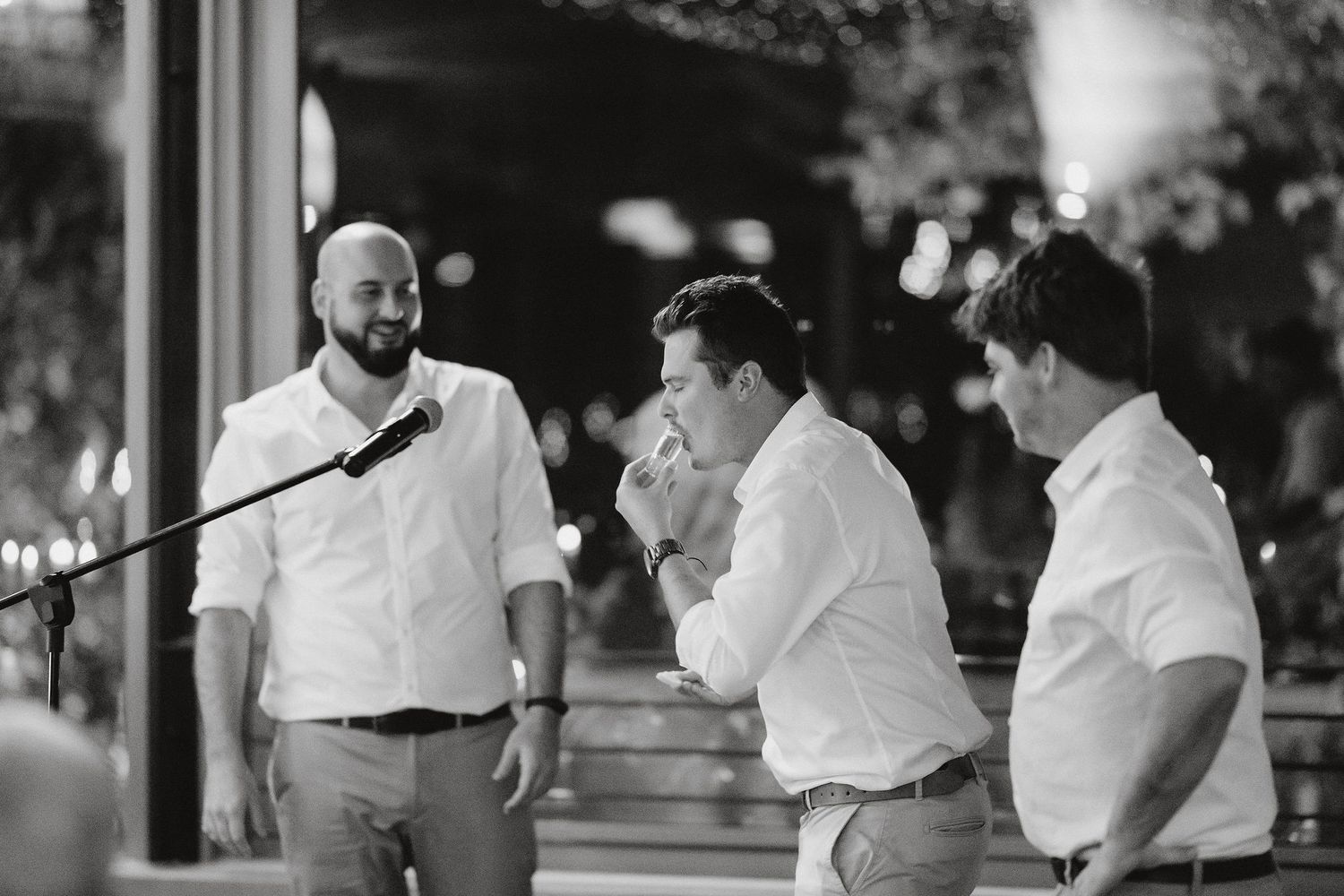 Three men in white shirts sharing a candid moment at an outdoor evening event, captured in black and white photography.