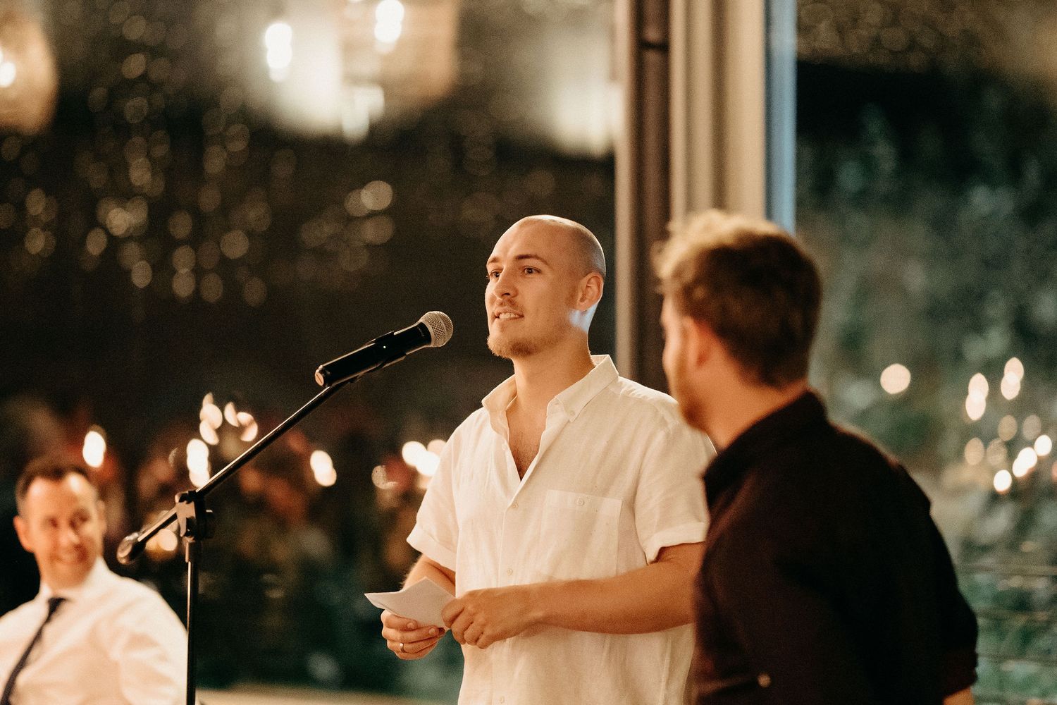 A speaker delivers a wedding speech at an outdoor evening reception with bokeh lights in the background.