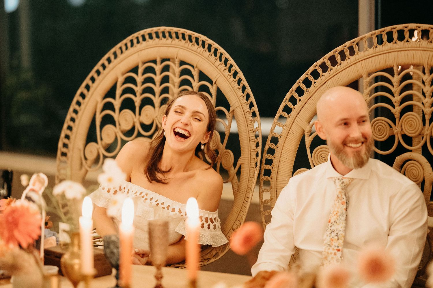 A candid moment of laughter during an evening reception with peacock chairs and glowing candles on the table.