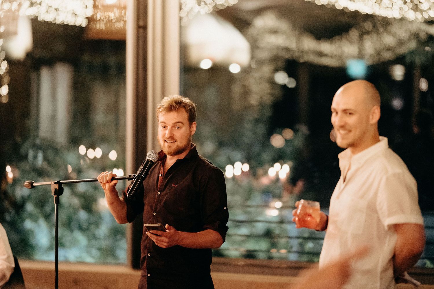Two people stand at a microphone during an evening event with twinkling lights in the background.