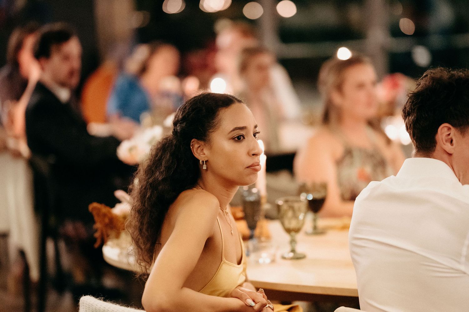 Elegant dinner party guest in golden dress sits thoughtfully at table during evening reception.