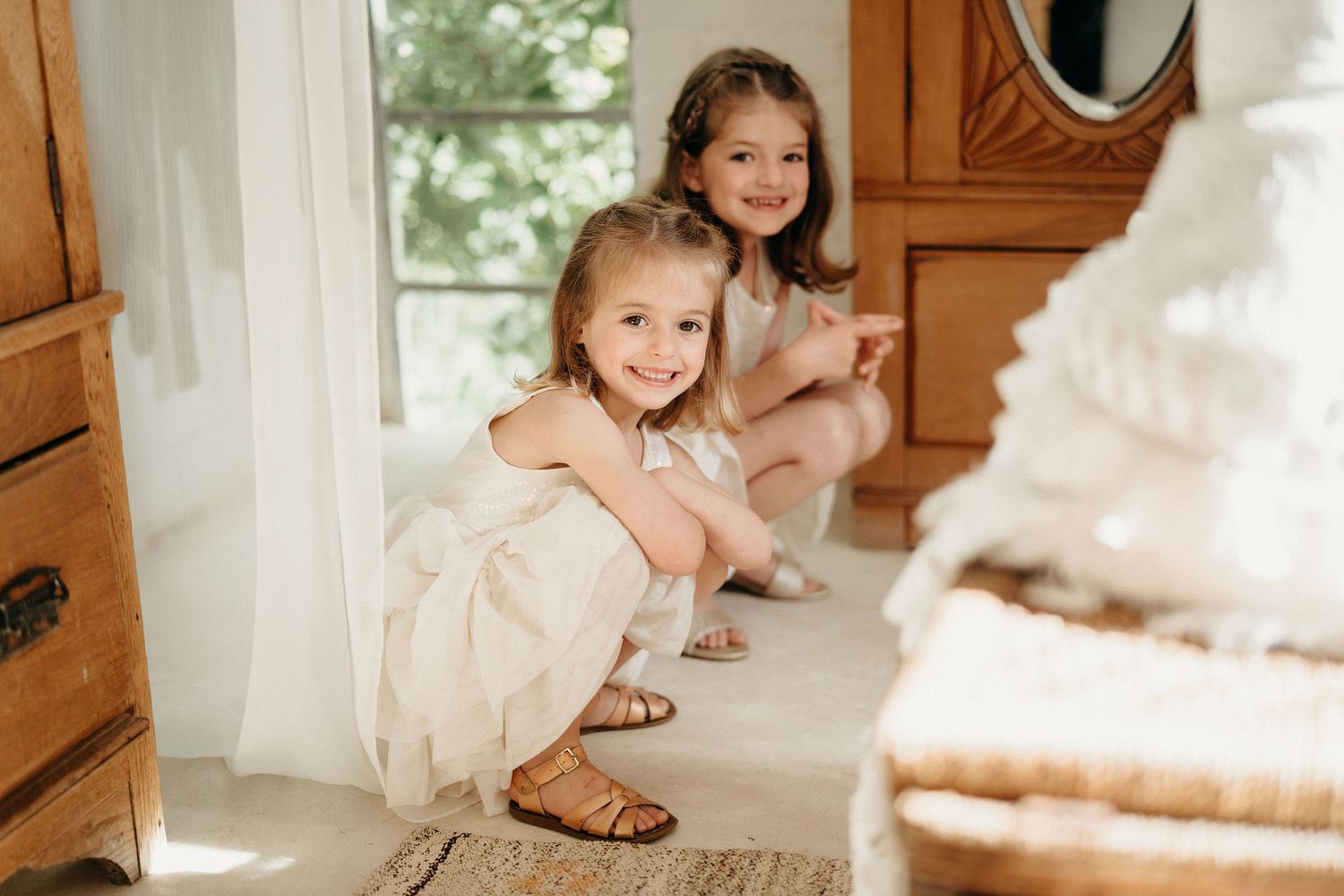 Two children in white dresses sit on a wooden floor and smile while looking up at something off camera.