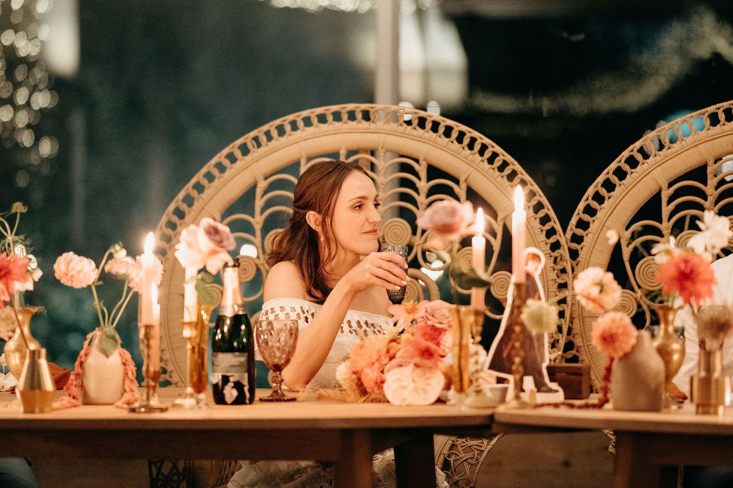 Two guests share a candlelit moment at an elegant reception with decorative peacock chairs and string lights.