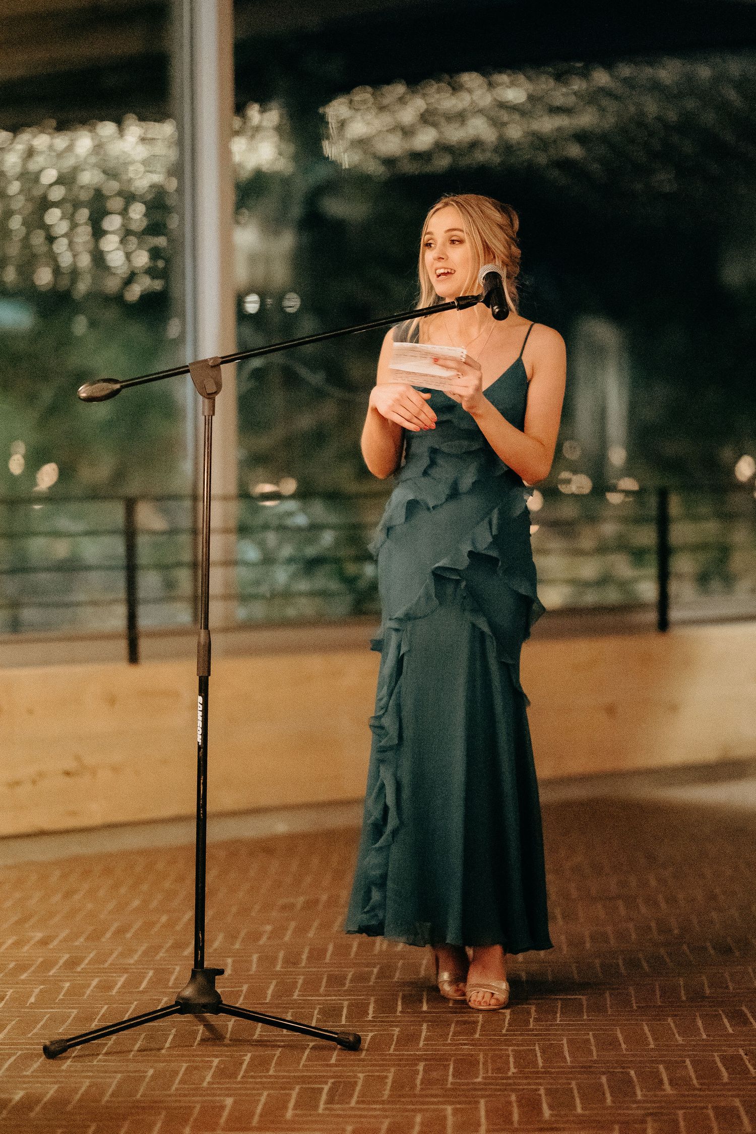 A person wearing an elegant dark teal gown stands at a microphone with twinkling lights in the background.