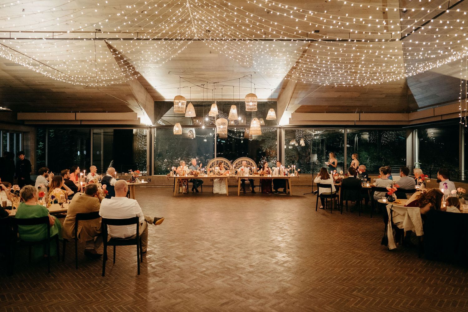 Indoor wedding reception venue with warm lighting, string lights overhead, and guests seated at long tables on a wood dance floor.