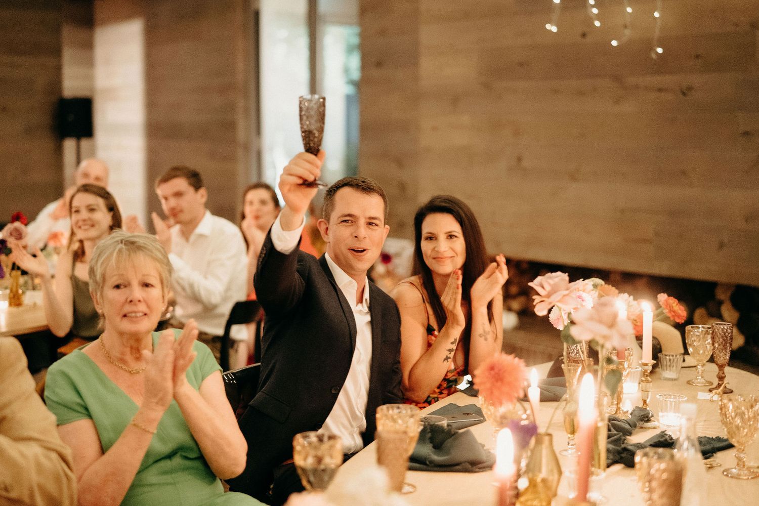 A group of guests celebrating at an elegant dinner party with raised glasses and candlelight in a wooden-paneled room.